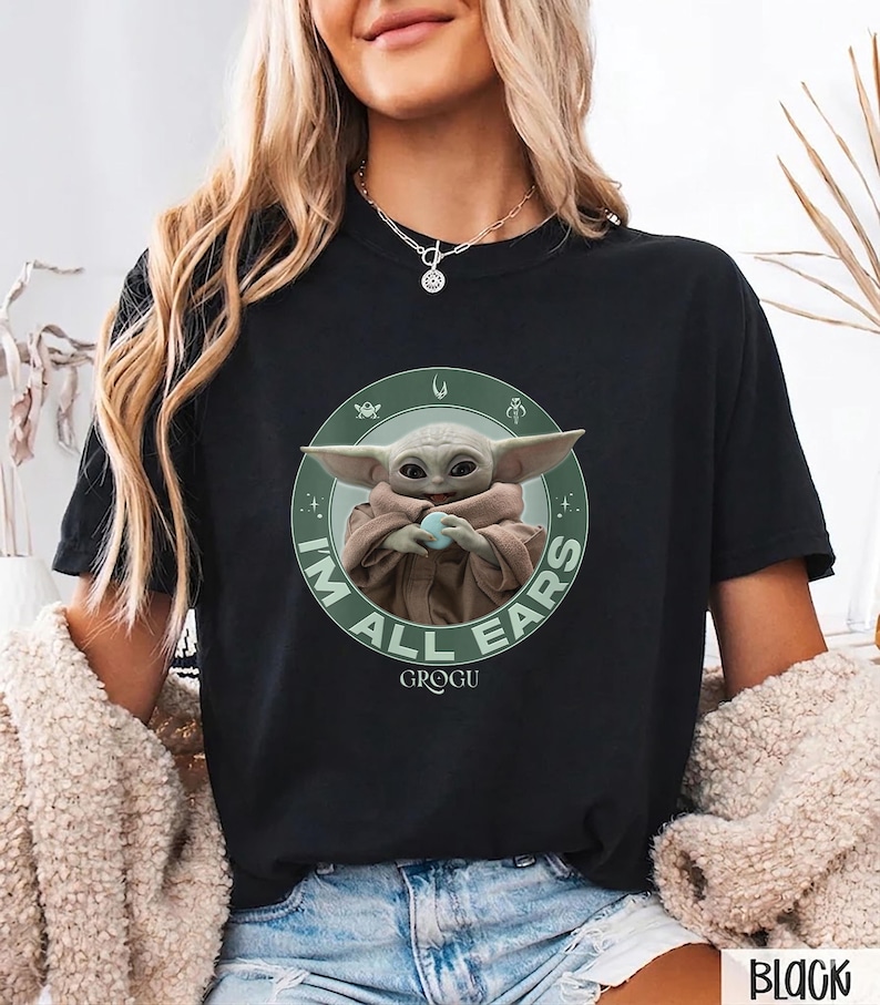 May include: Black t-shirt featuring a graphic of Grogu, also known as Baby Yoda, from The Mandalorian. The design includes the text "I'M ALL EARS" in green around Grogu. The shirt is a crew neck style.