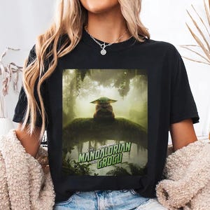 K&ouml;nnte beinhalten: Schwarzes T-Shirt mit einer Grafik von The Mandalorian und Grogu. Das Design zeigt Grogu in einer Waldszene. Der Text "The Mandalorian and Grogu" ist in Gr&uuml;n und Wei&szlig;. Das Shirt ist ein l&auml;ssiger Stil.