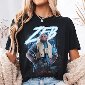 May include: Black t-shirt with a graphic of Zeb from the Mandalorian series. The design includes the name "ZEB" in blue lightning, the text "MANDALORIAN GROGU", and "CARAZED ORELIOS". The shirt is a crew neck.