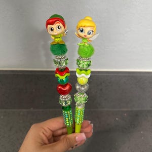 May include: Two decorative pens featuring character toppers of a boy in a green hat and a fairy with blonde hair. Each pen is adorned with a green fuzzy pom-pom, stacked faceted beads in shades of green, red, and silver, and a sparkling barrel.