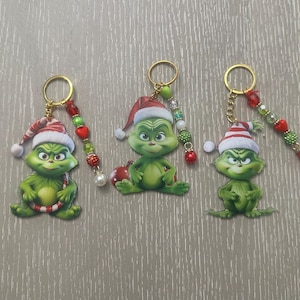May include: Three keychains featuring a green character wearing a red and white Santa hat. Each keychain includes a gold-tone ring and a dangling charm strand composed of red, green, and white beads in various textures, shapes, and sizes.