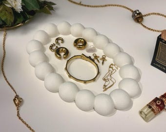 May include: A collection of gold-toned jewelry, including a bracelet, earrings, and a necklace, arranged on a white, oval-shaped tray. The tray is surrounded by white, spherical decorations. A small bottle with a red and gold design is also visible.