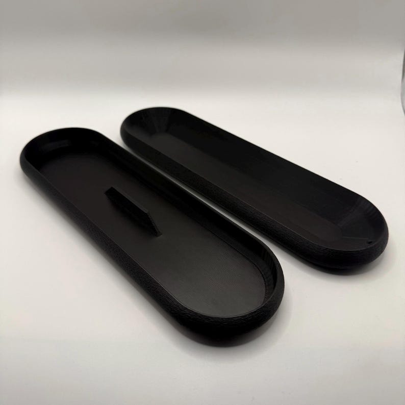 May include: Two black, elongated trays with rounded ends. One tray has a raised, rectangular divider in the center. The trays are made of a matte material and are set against a white background. The trays appear to be empty.