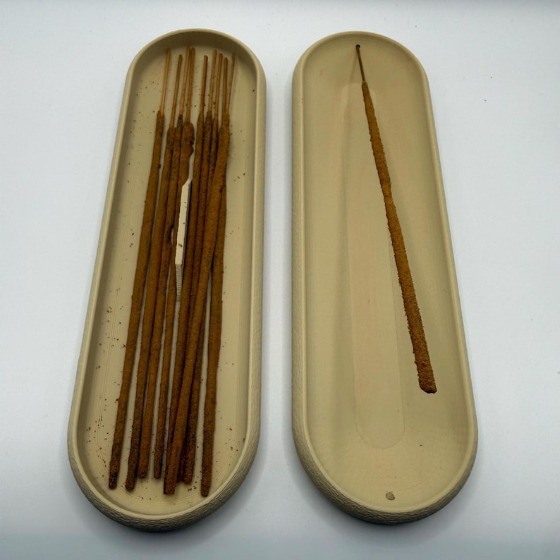 May include: Two beige, oval-shaped incense holders. One holder contains multiple brown incense sticks, while the other holds a single stick. The holders are placed on a white surface.
