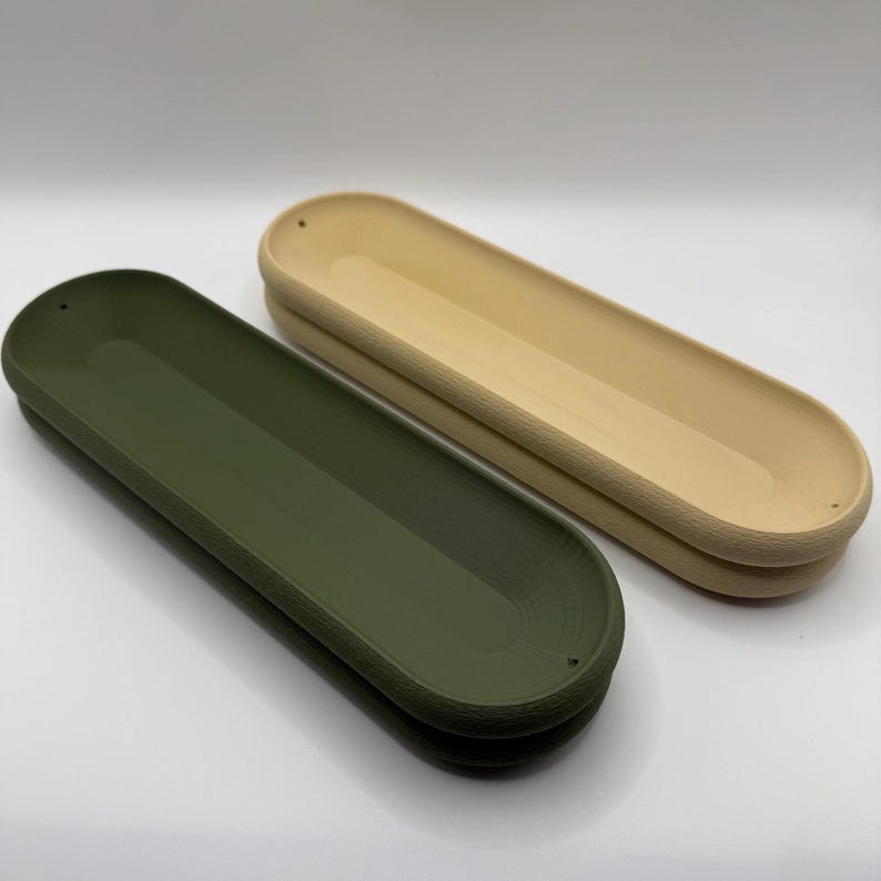 May include: Two long, oval-shaped trays. One is olive green, the other is a light beige. Both have a textured, ridged border and a smooth, recessed interior. The trays are designed with small holes at each end.