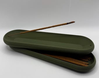 May include: An olive green incense holder with a textured surface. A lit incense stick is placed on top, with smoke rising. The holder contains several unlit incense sticks. The holder is oval-shaped and made of a solid material.