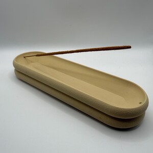 May include: A beige ceramic incense holder with a layered design. The holder is oval-shaped and features a groove to catch ash. A lit incense stick is placed in the holder, emitting a thin trail of smoke. The background is plain white.