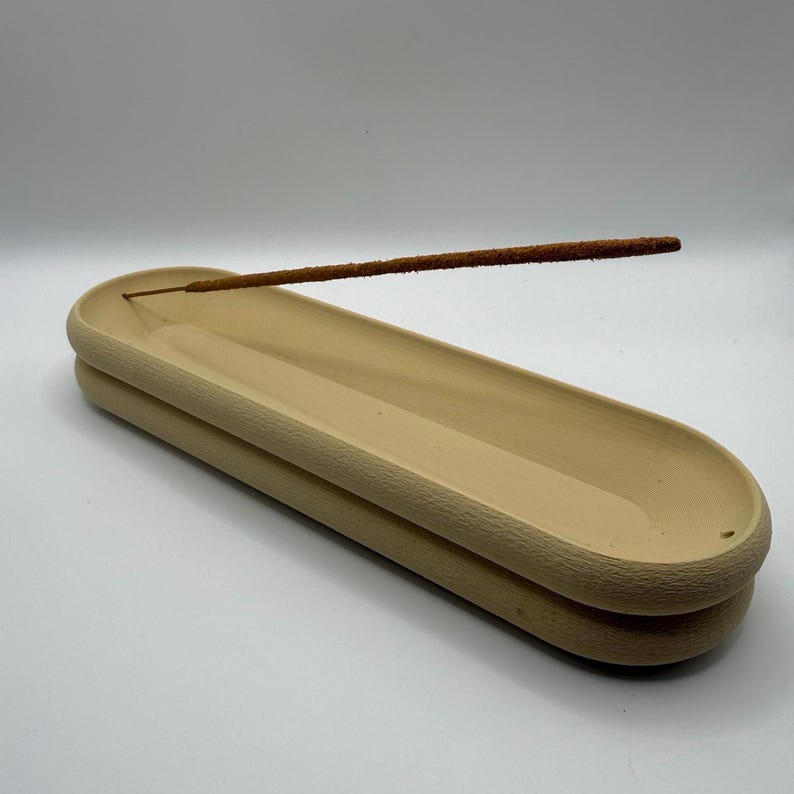 May include: A beige ceramic incense holder with a layered design. The holder is oval-shaped and features a groove to catch ash. A lit incense stick is placed in the holder, emitting a thin trail of smoke. The background is plain white.