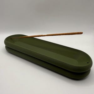 May include: An olive green incense holder with a textured surface. A brown incense stick rests in the holder. The holder is oval-shaped and has a stacked design. The background is white.