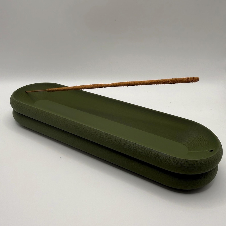 May include: An olive green incense holder with a textured surface. A brown incense stick rests in the holder. The holder is oval-shaped and has a stacked design. The background is white.