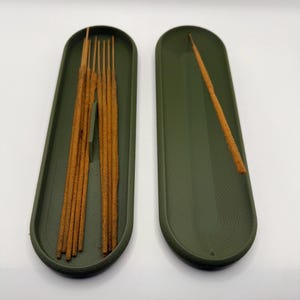 May include: Two olive green incense holders, each with a rounded rectangular shape. One holder contains several brown incense sticks, while the other holds a single stick. The holders are placed on a white surface.