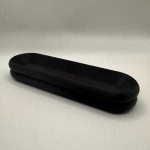May include: A black, elongated wooden tray with a rounded, oval shape. The tray is made of two stacked pieces, creating a layered effect. The surface is smooth and the color is a deep, solid black. The tray is designed for decorative or functional use.