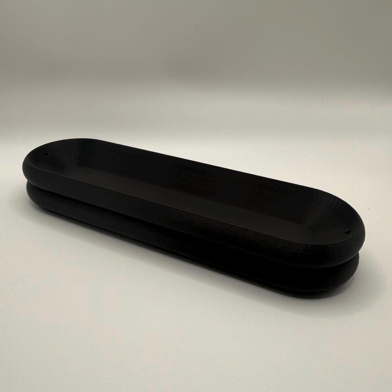 May include: A black, elongated wooden tray with a rounded, oval shape. The tray is made of two stacked pieces, creating a layered effect. The surface is smooth and the color is a deep, solid black. The tray is designed for decorative or functional use.