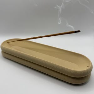May include: A beige ceramic incense holder with a textured rim, holding a lit incense stick. Wisps of smoke rise from the incense, creating a calming visual. The holder has a layered design, adding a touch of elegance to the aromatherapy accessory.
