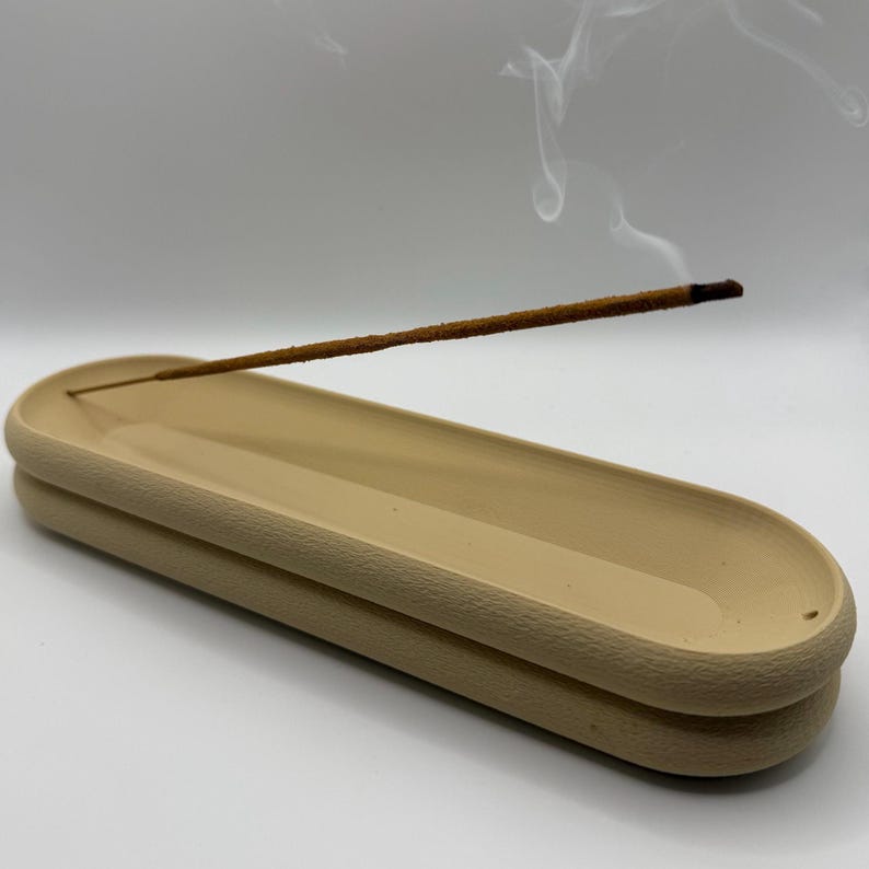 May include: A beige ceramic incense holder with a textured rim, holding a lit incense stick. Wisps of smoke rise from the incense, creating a calming visual. The holder has a layered design, adding a touch of elegance to the aromatherapy accessory.