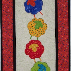 May include: A colorful quilted wall hanging with a row of five sheep, each with a different colored body and a different colored face. The sheep are arranged in a vertical line, with the top sheep at the top of the image and the bottom sheep at the bottom of the image. The sheep are all facing the right side of the image. The background of the image is a white fabric with a floral pattern.