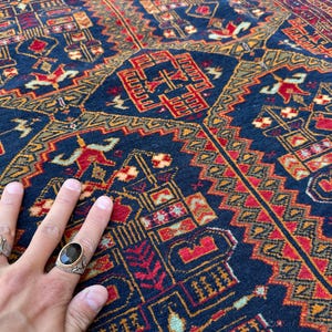 Könnte beinhalten: Nahaufnahme einer Hand auf einem gemusterten Teppich. Der Teppich hat einen tiefblauen Hintergrund mit komplizierten geometrischen Mustern in Rot, Gold und Braun. Die Hand trägt zwei Ringe, einen mit einem gelben Stein und einen mit einem schwarzen Stein.