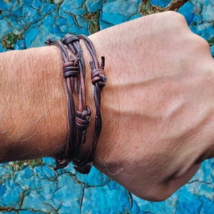 May include: A dark brown leather bracelet designed to resemble barbed wire, featuring multiple twisted strands and knotted sections that wrap around the wrist. The accessory has a weathered, textured appearance with distinct dark brown tones.