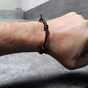 May include: A dark brown leather bracelet with a knotted design is worn on a person's wrist. The bracelet is wrapped around the wrist and secured with a knot. The background is a gray concrete surface.