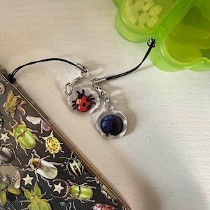 May include: Two clear acrylic charms with black cords. One charm features a red ladybug with black spots, and the other has a blue sphere design. The charms are attached to a phone case decorated with insect illustrations.