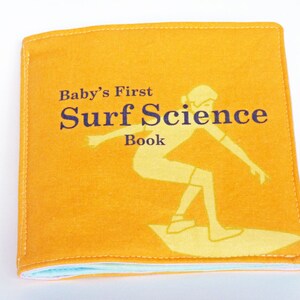 Puede incluir: Un libro de tela amarillo con el t&iacute;tulo "Baby's First Surf Science Book" y una ilustraci&oacute;n de una persona surfeando.