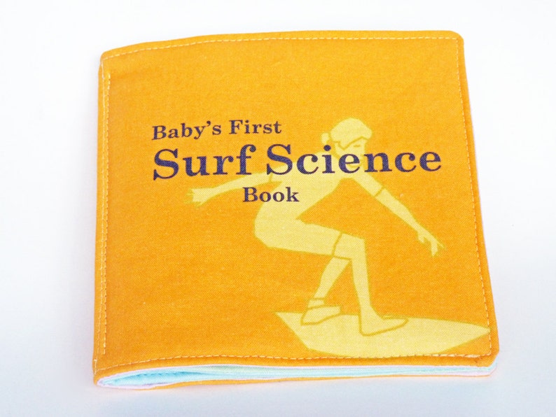 Puede incluir: Un libro de tela amarillo con el t&iacute;tulo "Baby's First Surf Science Book" y una ilustraci&oacute;n de una persona surfeando.