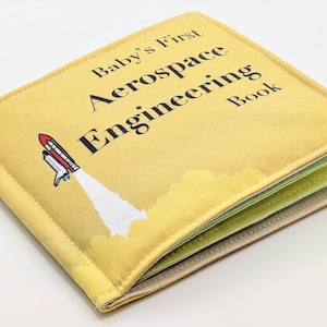 Puede incluir: Un libro de tela amarillo con el título "Baby's First Aerospace Engineering Book" y una ilustración de un cohete despegando.