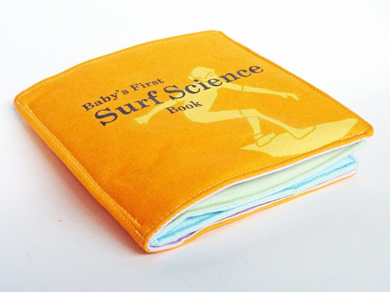 Puede incluir: Un libro amarillo para beb&eacute;s con el t&iacute;tulo "Baby's First Surf Science Book" y una silueta de una persona surfeando en una ola.