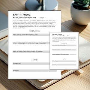 May include: Two white pages with the text "FAITH IN FOCUS" and prompts for reflection and action, with a smaller version of the same pages. The pages are on a light-colored leather notebook on a wooden surface.