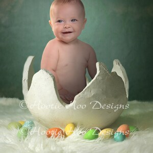 May include: A baby sitting inside a white cracked egg shell prop. The baby is wearing no clothes and is smiling. The egg shell is surrounded by colorful Easter eggs on a white fluffy surface.