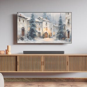 Peut inclure: Une peinture à l'aquarelle encadrée représente un village européen enneigé avec des bâtiments, des arbres à feuilles persistantes et un éclairage doux. L'œuvre est au-dessus d'un meuble en bois avec une barre de son.