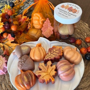 Puede incluir: Un conjunto de jabones con temática otoñal con forma de calabazas, hojas y bellotas. Los jabones son en tonos naranja, marrón y rosa, con detalles dorados. Algunos jabones están en un tarro transparente con tapa blanca.