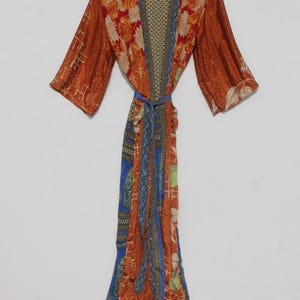 May include: A long, patterned robe in shades of orange, red, blue, and green, hanging on a wooden hanger. The robe features floral and geometric designs, with a matching tie belt. The sleeves are wide and the fabric appears lightweight.