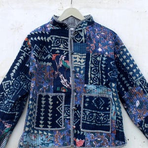 Puede incluir: Chaqueta acolchada azul con diseño patchwork, con patrones geométricos y florales. Tiene cuello alzado, mangas largas y dos bolsillos delanteros. Costuras blancas delinean los parches y bordes.