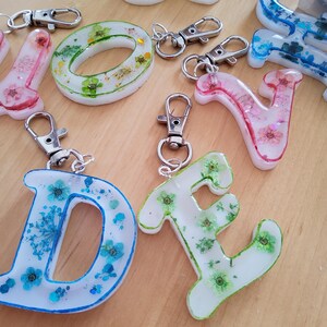 Könnte beinhalten: Verschiedene Schlüsselanhänger aus Kunstharz in Form von Buchstaben und einem 'O', jeweils mit getrockneten Blumen eingebettet. Die Buchstaben sind in verschiedenen Farben, darunter Rosa, Blau und Grün, und sind an silberfarbenen Karabinerverschlüssen befestigt.