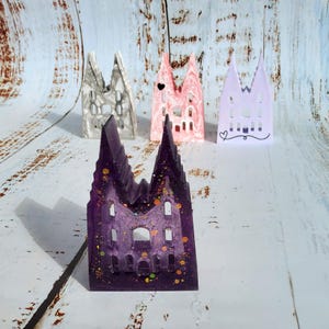 May include: A selection of miniature cathedral-shaped resin sculptures in various colours. The front sculpture is a deep purple with gold glitter. Other sculptures are white, pink, and lavender. Each has architectural details like arched windows and spires.