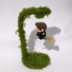 Puede incluir: Una pequeña figura de ganchillo con cabello castaño y traje negro, montada sobre un alambre fino para parecer que vuela sobre una escoba color canela. La figura se sitúa junto a una estructura curva cubierta de musgo sobre una base circular.