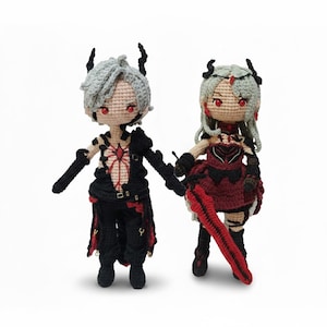 May include: Two handmade crochet dolls, one male and one female, stand side-by-side. The male doll wears a black outfit with red accents, while the female doll is in a red and black dress. Both dolls have gray hair, red eyes, and small horns.