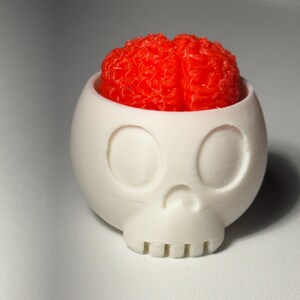 Può includere: Un contenitore bianco a forma di teschio con un cervello rosso all'interno. Il contenitore ha orbite oculari, una cavità nasale e denti. Il cervello è di colore rosso vivo e ha una superficie strutturata. L'articolo è probabilmente un pezzo decorativo o un oggetto di novità.