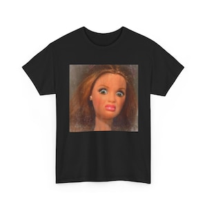 Può includere: T-shirt nero con un'immagine in primo piano del volto di una bambola vintage con un'espressione sorpresa. La bambola ha capelli castani, occhi grandi e una bocca rosa. L'immagine è centrata sulla maglietta, creando un design audace.