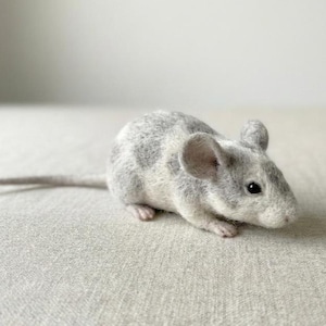 Puede incluir: Un ratón de fieltro hecho a mano en tonos grises y blancos. El ratón tiene una cola larga, pequeños pies rosados y orejas grandes y redondeadas. El cuerpo está moteado con manchas grises más oscuras. El ratón está sobre una superficie gris claro.