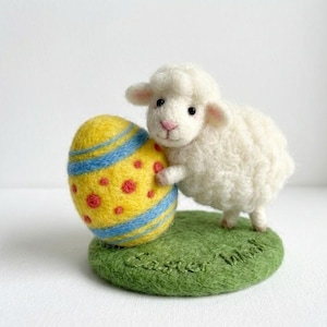 May include: A white, fluffy sheep figurine stands next to a yellow Easter egg decorated with blue stripes and red dots. The sheep is on a green felt base with the words "Easter Wishes" embroidered on it. The figurine is a festive Easter decoration.