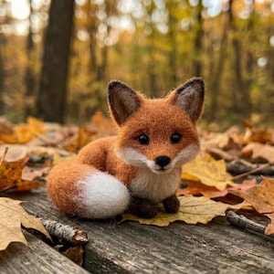 May include: A handcrafted, orange and white fox figurine with black eyes and ears. The fox is lying on a wooden surface covered with fallen autumn leaves. The background features a blurred forest scene.