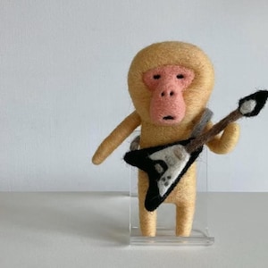 Puede incluir: Figura de mono hecha a mano en fieltro, con cara rosa y cuerpo marrón claro, sosteniendo una guitarra eléctrica negra y blanca. El mono está sobre un soporte de acrílico transparente sobre fondo blanco.