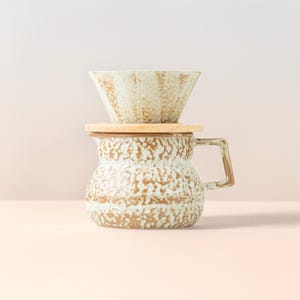 Può includere: Un set per la preparazione del caffè a versamento in ceramica. Il set include una caffettiera strutturata, marrone chiaro e bianca, con manico, coperchio in legno e cono versatore abbinato. Il design ha un'estetica rustica e artigianale.