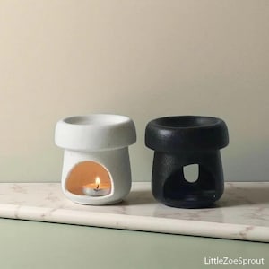 May include: Two ceramic oil warmers, one white and one black, sit side-by-side. The white warmer has a lit tea light inside, visible through a curved opening. The black warmer has a similar shape but no visible light. The warmers are on a marble surface.