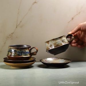 May include: Handmade ceramic teacups and saucers. The teacups have a unique, textured design with brown, blue, and cream tones. One cup is being held, while another sits on a saucer. The set includes additional saucers, showcasing a rustic, artisanal aesthetic.