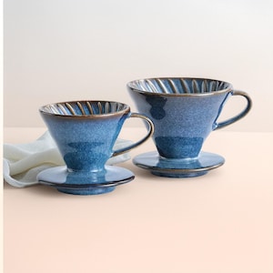 May include: Two blue ceramic pour-over coffee makers. Each has a cone shape, a handle, and a small, round base. The rims are a darker brown color. The coffee makers are on a light pink surface.