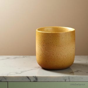 Op de afbeelding: Een cilindrische, mosterdgele keramische pot met een textuur. De pot staat op een marmeren aanrecht. De pot heeft een afgeronde bodem en een brede opening. De achtergrond is neutraal.