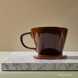 Puede incluir: Un vertedor de café de cerámica marrón con asa y base circular. El vertedor de café es brillante y se asienta sobre una superficie de mármol blanco y gris. El fondo es de color beige liso.